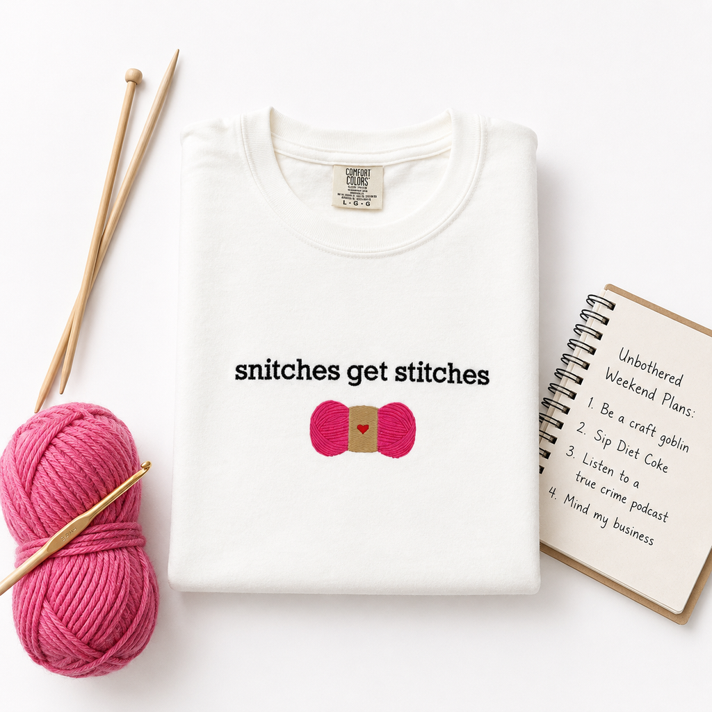 A folded white t-shirt with the text 'snitches get stitches' and a pink skein of yarn embroidered design, styled in a flat lay with knitting needles, pink yarn, and a notebook on a white background.